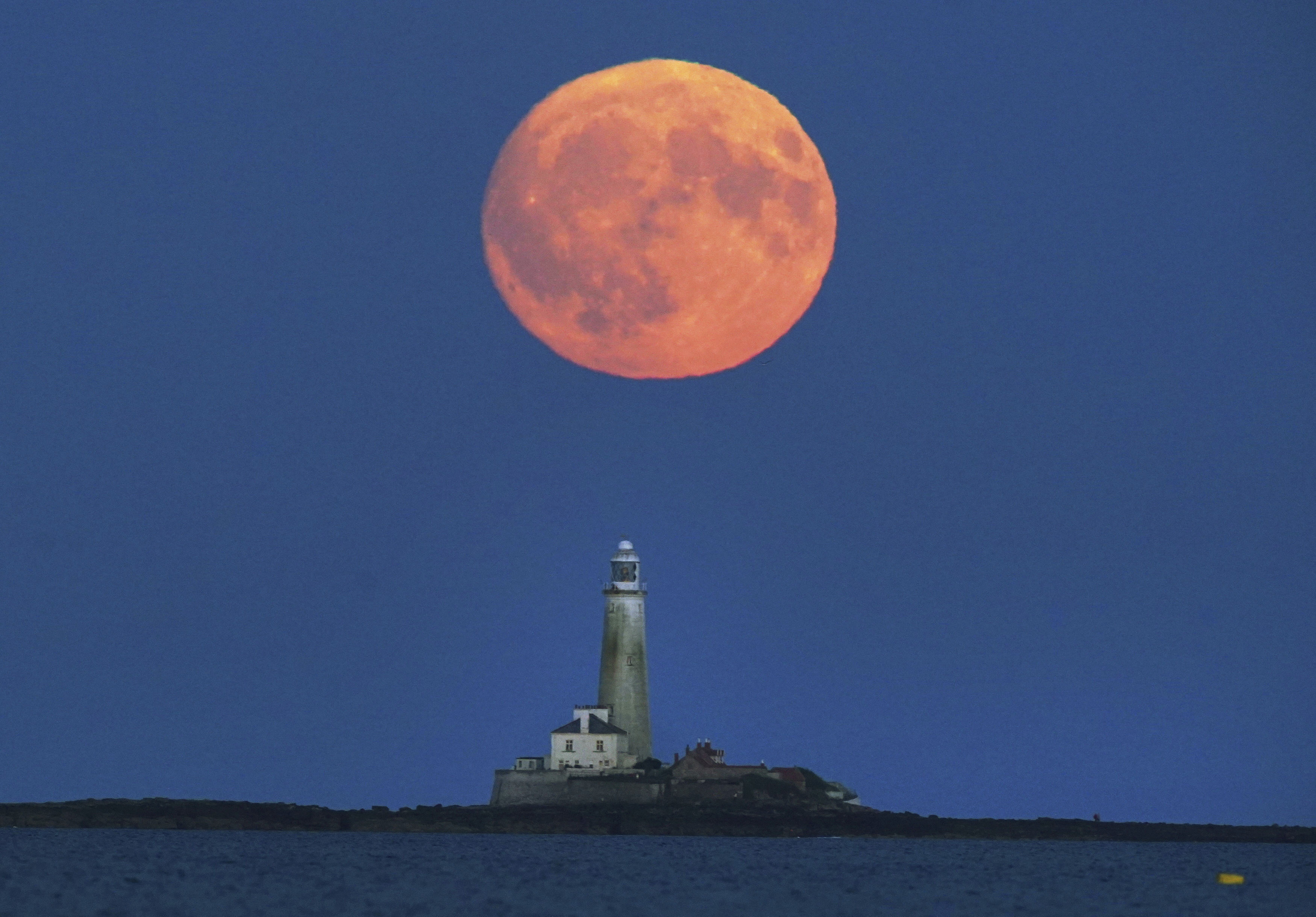 A large orange moon rises over a lighthouse sitting on a shallow rise.