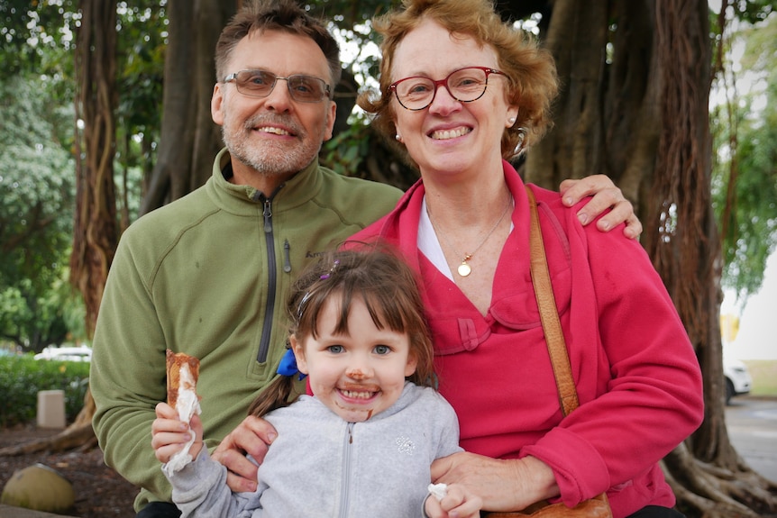 A man and a woman in jumpers with their arms around a young girl holding an ice cream cone with chocolate on her face