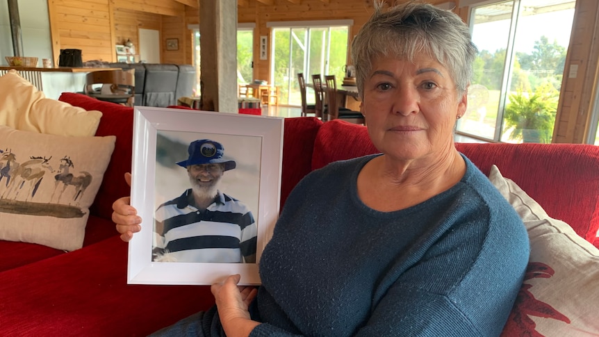 A middle-aged white woman with short grey hair. She is sitting down, wearing a blue sweater, holding a photo of her dead husband