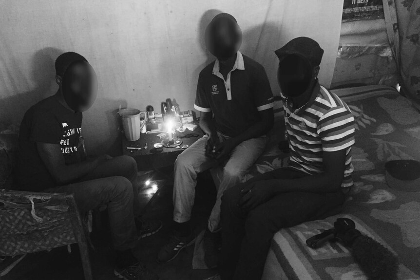 Three men sit close together around a table with a candle and drug utensils.