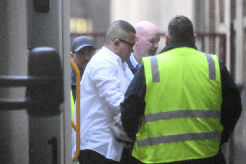 A man with short dark hair and a white shirt is escorted by a man in a high vis vest while in custody.