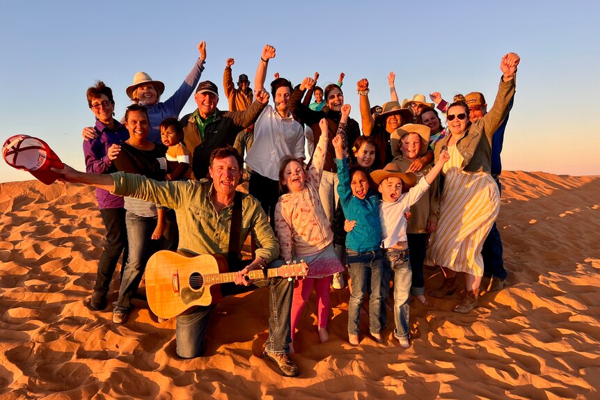 Group of people laughing, with arms in the air standing on a big sand dune at sunset. 