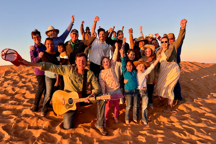 Group of people laughing, with arms in the air standing on a big sand dune at sunset. 