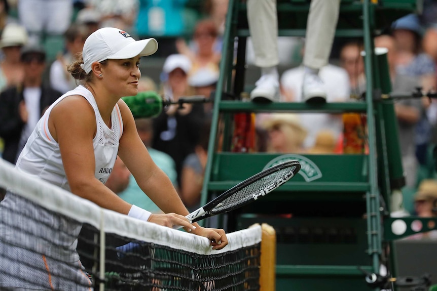 Ash Barty leans on the net holding her racquet with a smile on her face