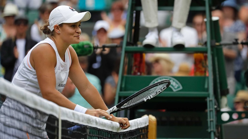 Ash Barty leans on the net holding her racquet with a smile on her face