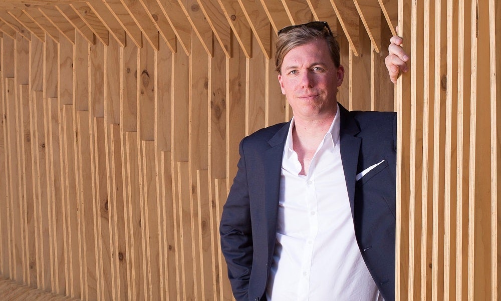 A man with blonde hair and sunglasses standing in front of a wall in a suit jacket