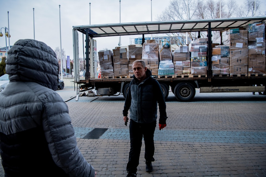 Two men in black puffer coats stand next to a truck stacked full of cardboard 