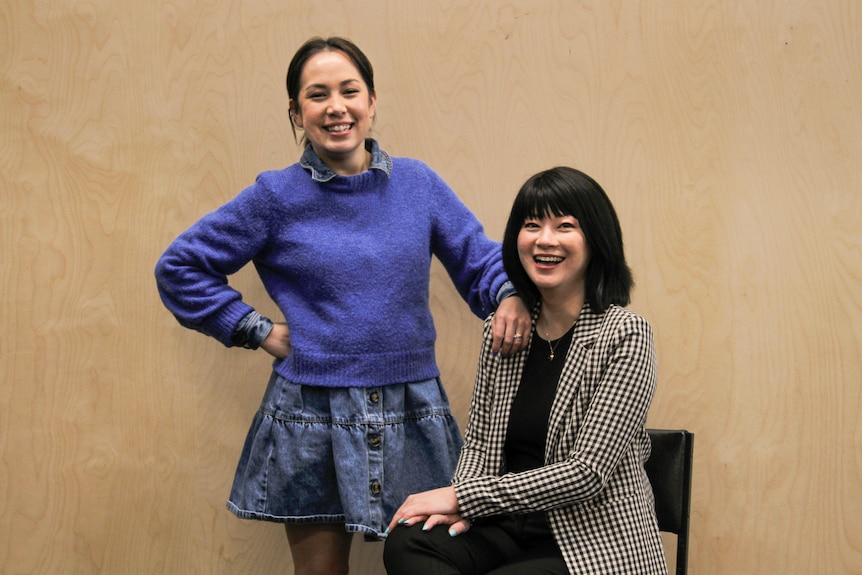 Two Asian Australian women in their 30s smile brightly, one standing with a hand on her hip behind the other, who is seated