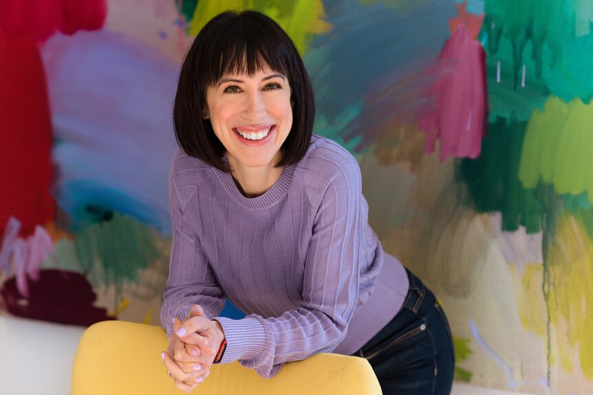 Amantha Imber smiles while leaning on chair. She's standing against a colourful background and wearing a purple blouse.