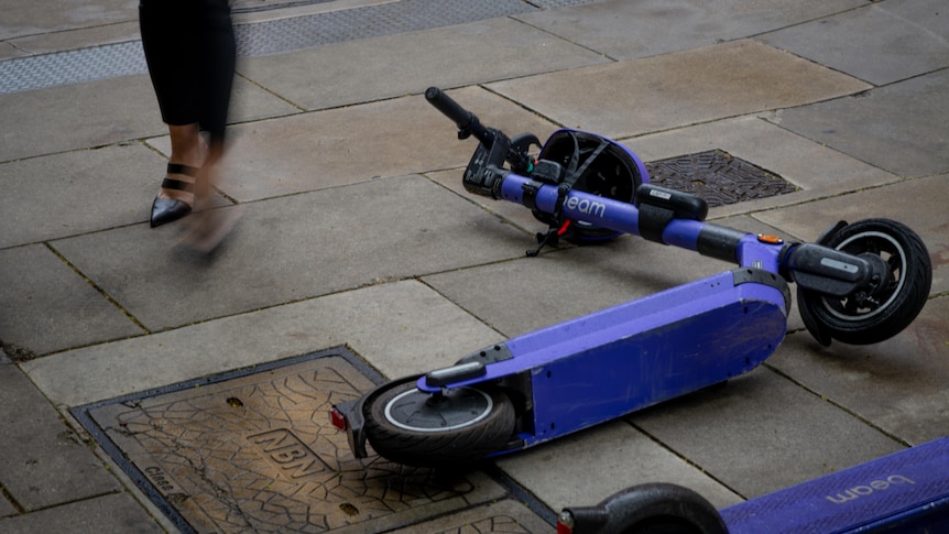 A purple e-scooter on its side on a foothpath, with someone's legs walking by