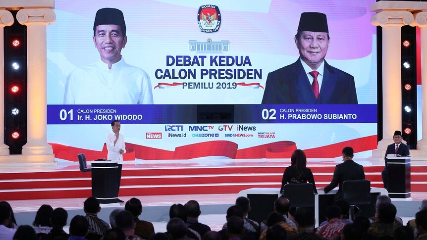 Two men debate on a large stage on which a screen displays their images and headshots.