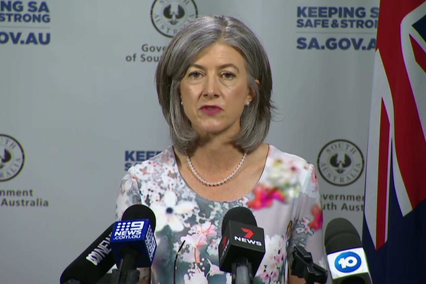 A woman with grey hair wearing a floral dress standing in front of a board with SA Government logos
