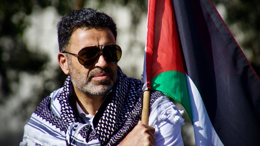 Man sits holding a Palestinian flag wearing sunglasses. 