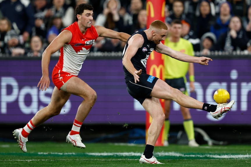A Carlton AFL player tries to hook the ball towards goal with his right boot as a Swans defender trails.