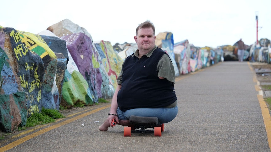 A man with no legs and one functional arm sits on a skateboard on the breakwall path in front of painted rocks.