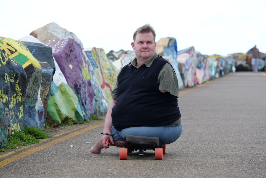 A man with no legs and one functional arm sits on a skateboard on the breakwall path in front of painted rocks.