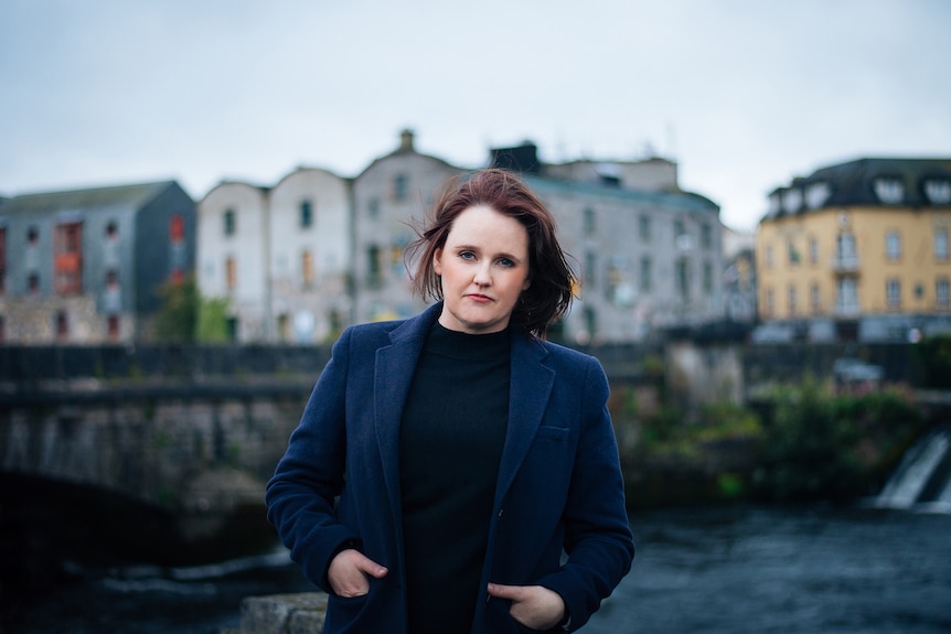 Portrait of Irish crime writer Dervla McTiernan, standing in front of a bridge and river