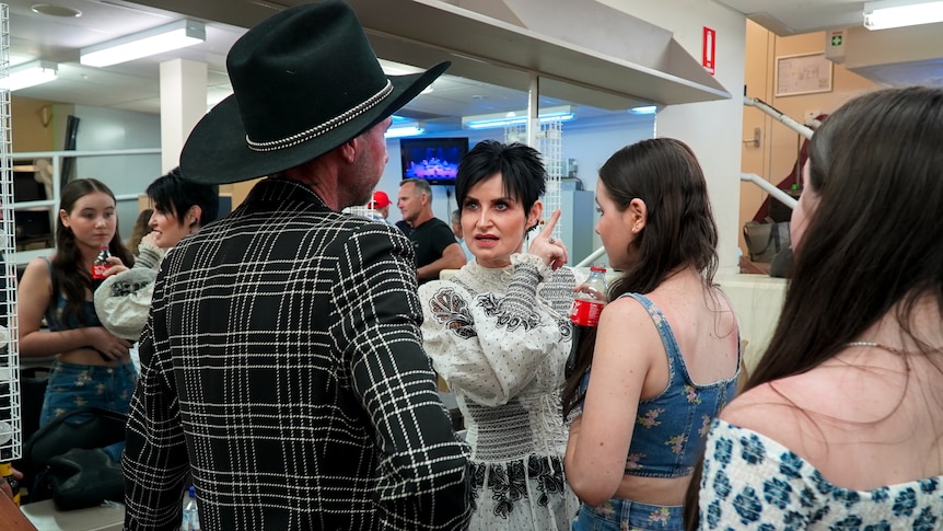 A woman talks to a man in a large felt hat while two teen-aged girls look on