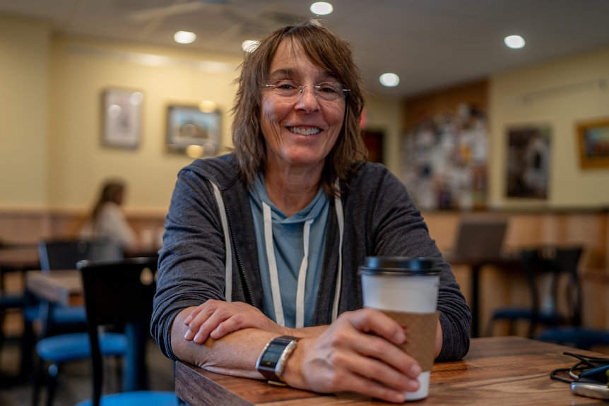 A woman in grey hoodie holds a coffee in a cafe and smiles at the camera