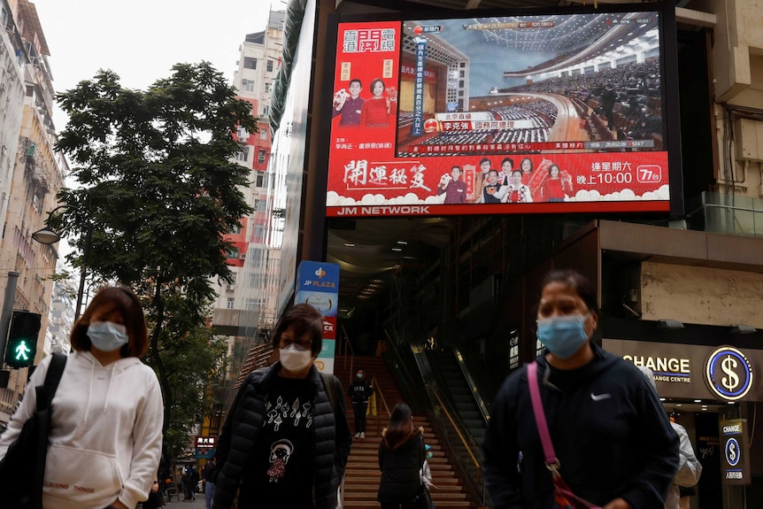 People wearing masks walk around a street below a big screen television airing the NPC.