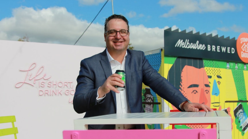 A man in a suit stands at an outdoor bar table with a beer, surrounded by colourful chairs and food and wine vans.