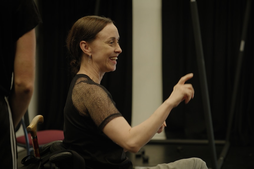 A white woman in her 50s with mousey brown hair, wearing a sheer black shirt, laughs while directing dancers in a studio.