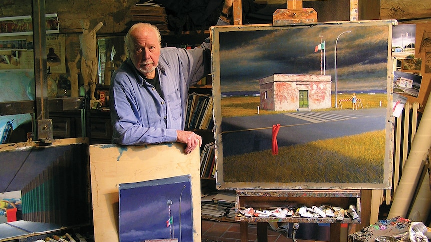 Balding older white man in blue shirt leans on canvas beside an art easel with a painting of a cordoned off street and building