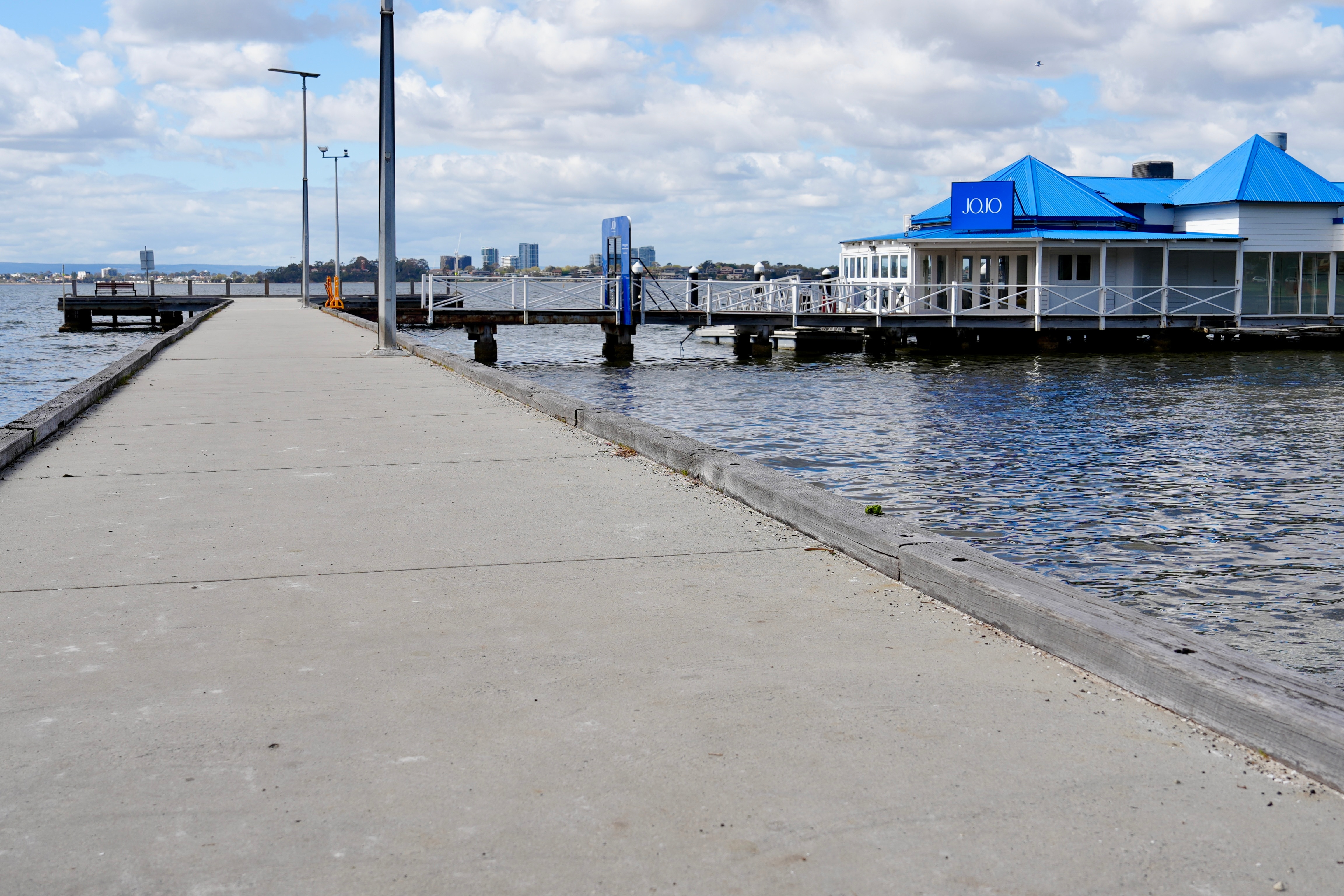 A concrete jetty in a river that leads to a white building with a bright blue roof and a sign that reads JoJo's.