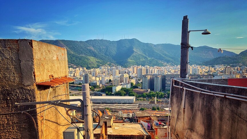 A panorama shot shows a favela in the foreground looking onto high-rise buildings with green mountains rising in the background