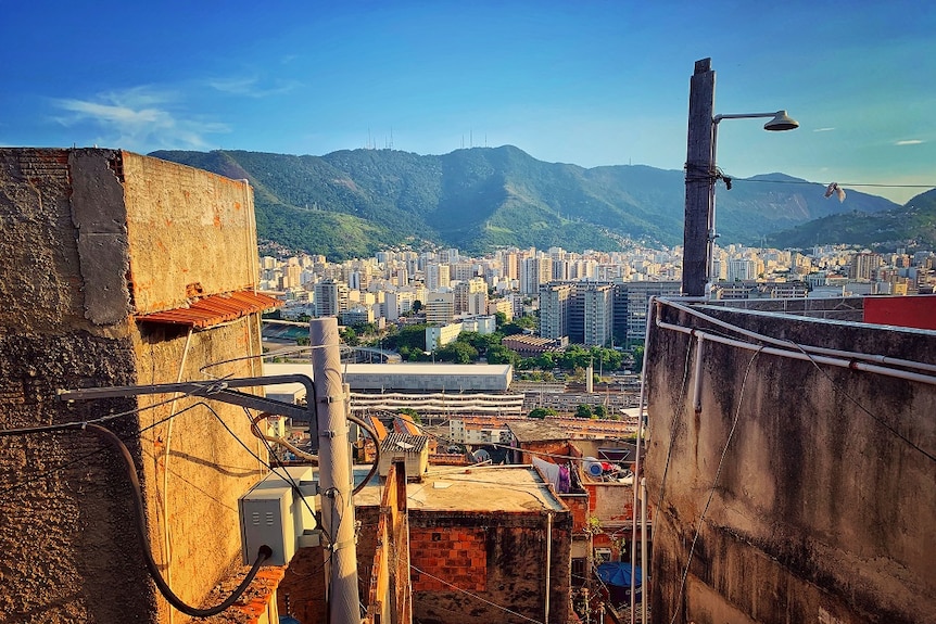 A panorama shot shows a favela in the foreground looking onto high-rise buildings with green mountains rising in the background