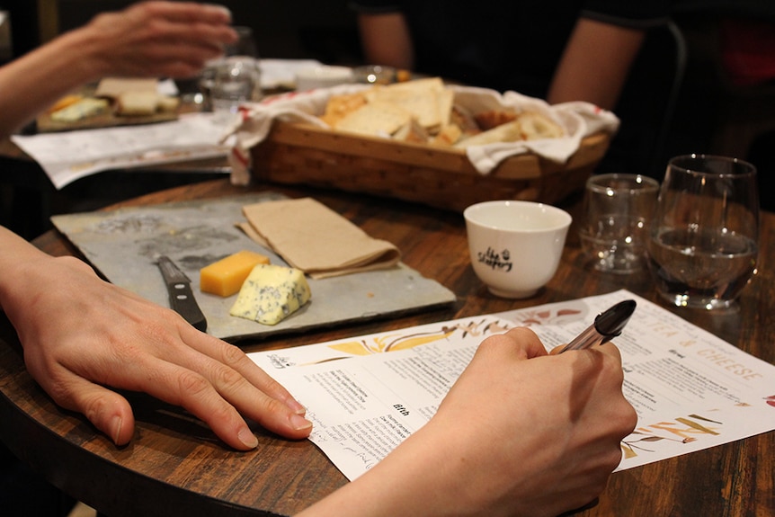 A woman's hands writing notes on a sheet of paper with cheeseboards, tea and baskets of bread and crackers in the background.