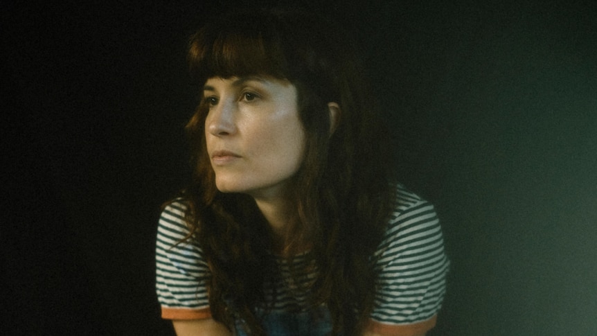 woman with dark hair sits in a dark room and stares into the distance