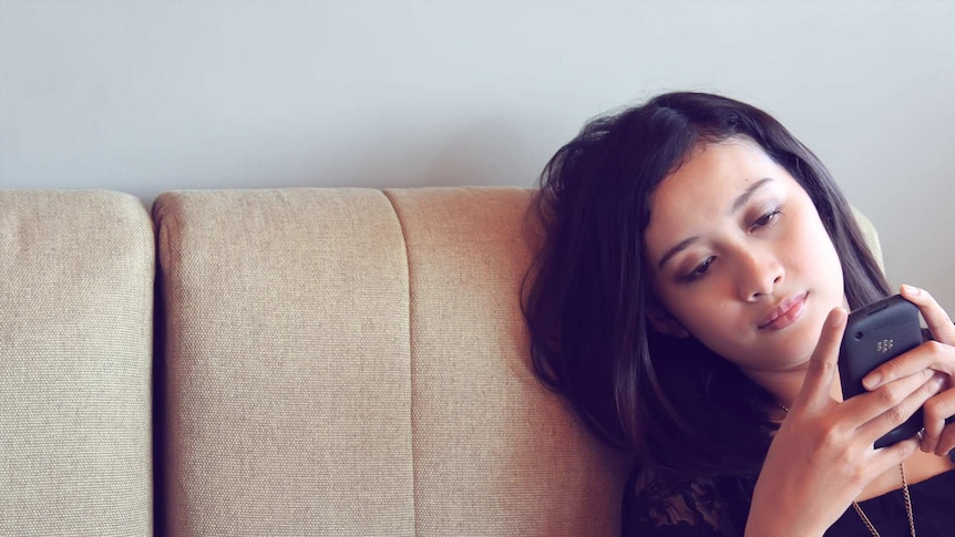 Young woman sitting on a beige couch looking at her phone and appearing lonely