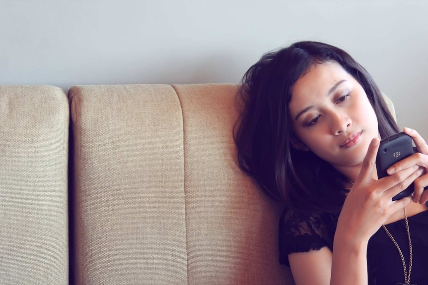 Young woman sitting on a beige couch looking at her phone to depict loneliness.