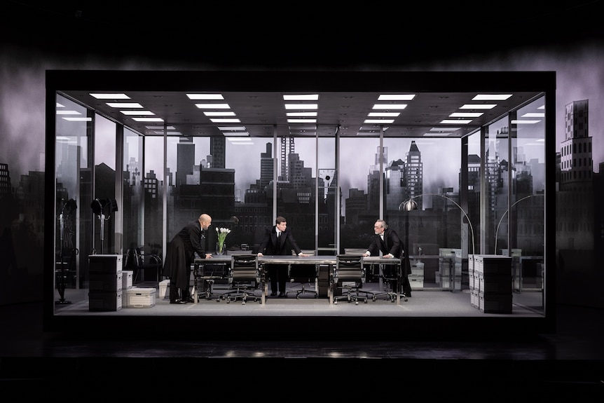 A scene of a stage featuring three male actors around a table inside a glass box with the New York skyline behind