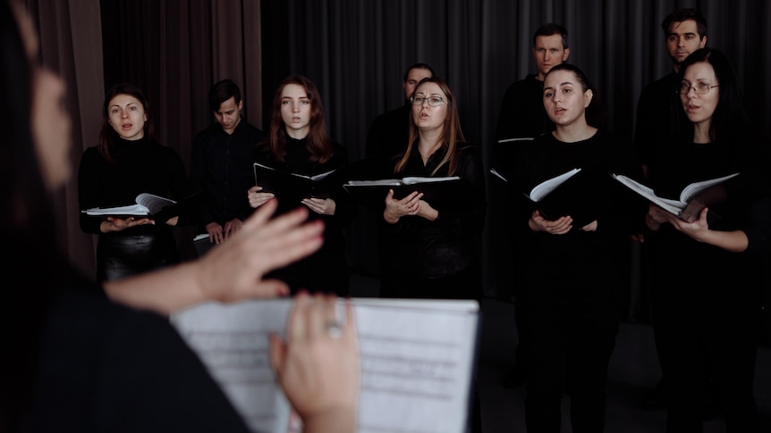 A choir singing while holding music notation. The conductor is at the front of the choir and you can see over their 