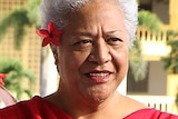 Fiame Naomi Mata'afa, with short grey hair, wearing a red dress, with a red flower in her hair.