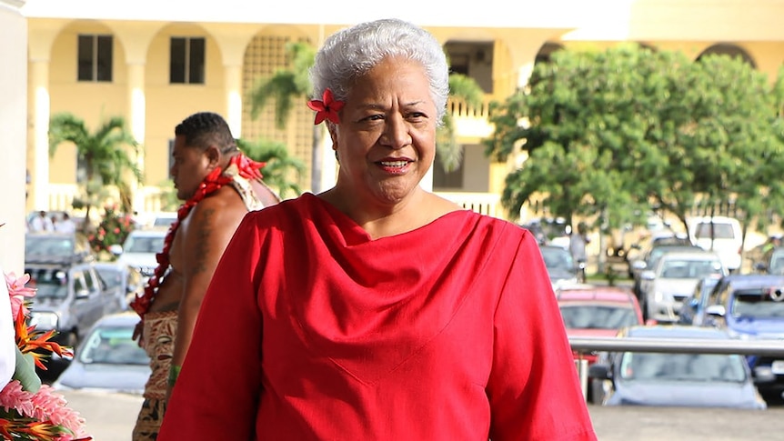 Fiame Naomi Mata'afa, with short grey hair, wearing a red dress, with a red flower in her hair.