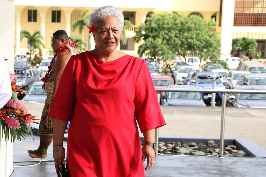 Fiame Naomi Mata'afa, with short grey hair, wearing a red dress, with a red flower in her hair.