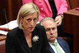 Katy Gallagher with a serious expression stands in Parliament in a black suit with a flower on her lapel