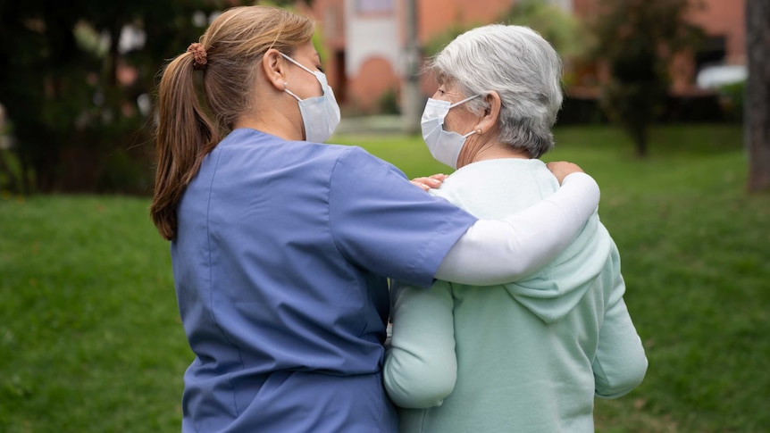 Caregiver walking at the park with a senior woman while wearing facemasks.