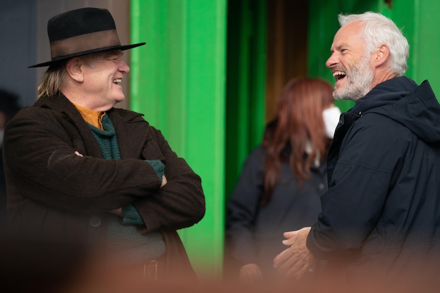 Two older men in dark coats laugh on a film set.