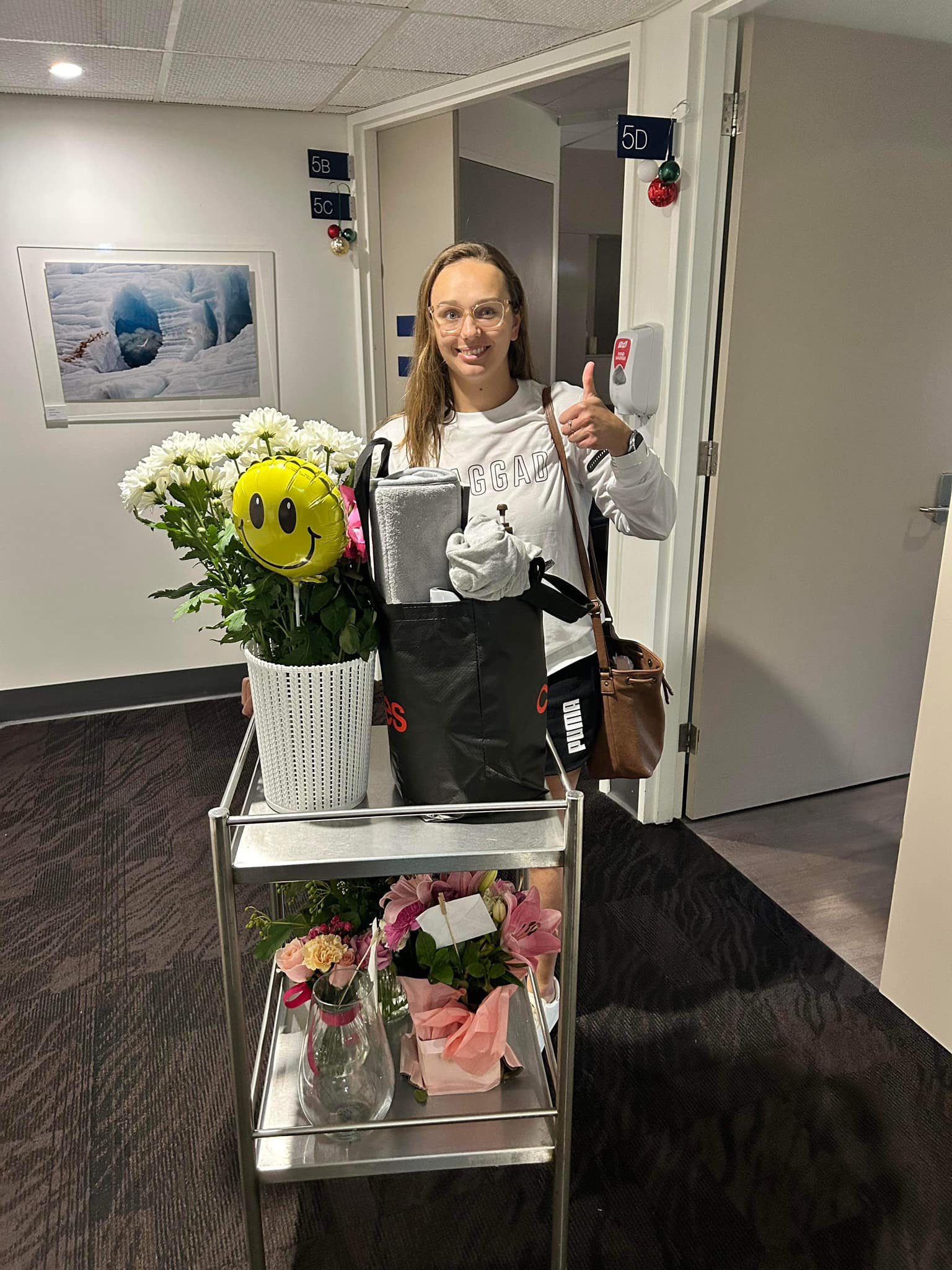 a young woman stands behind a medical trolley with flowers and gives thumbs up