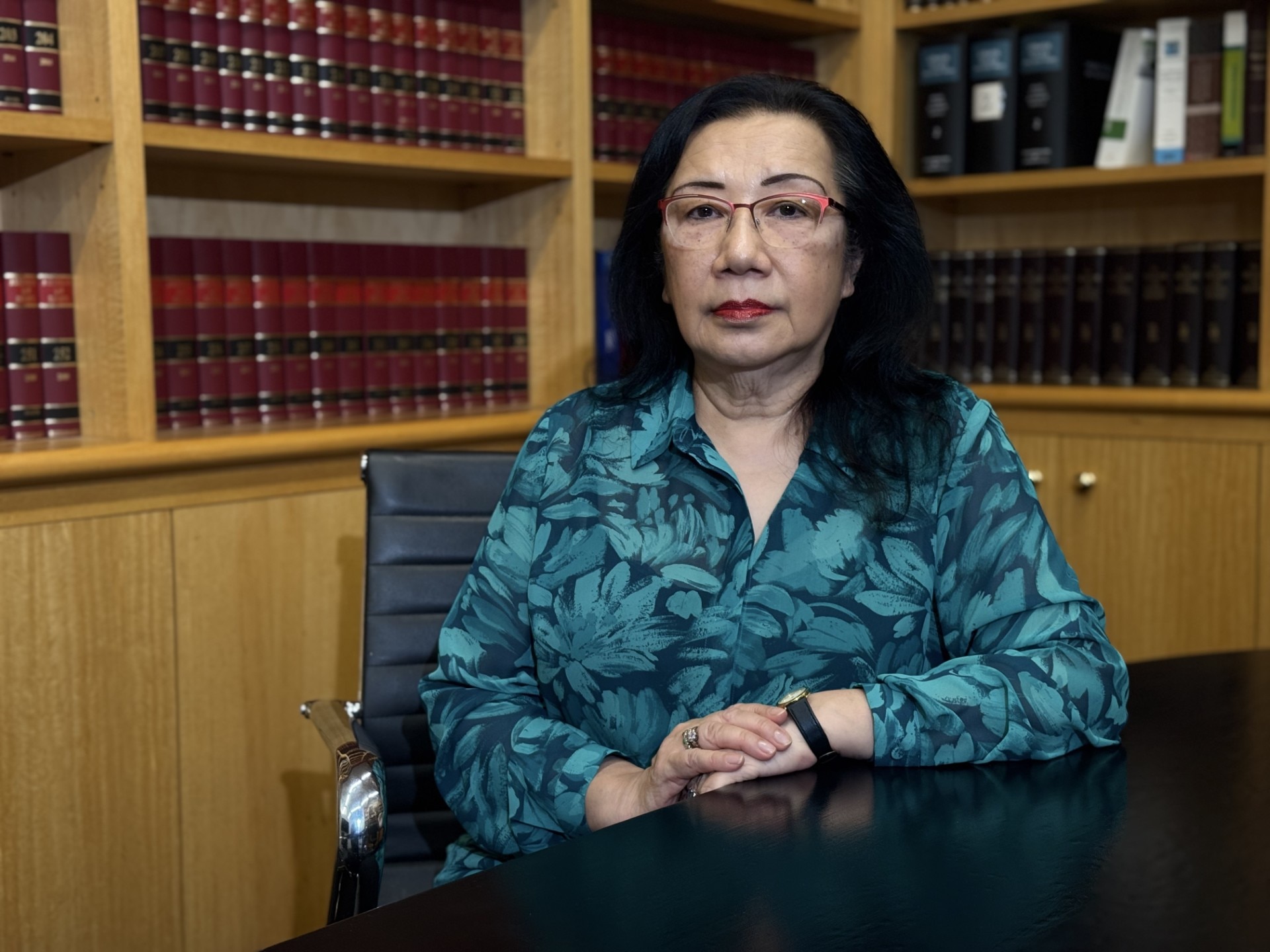 A profile shot of a woman in a blue flower-patterned shirt, glasses and black hair, seated at a table with rows of books behi
