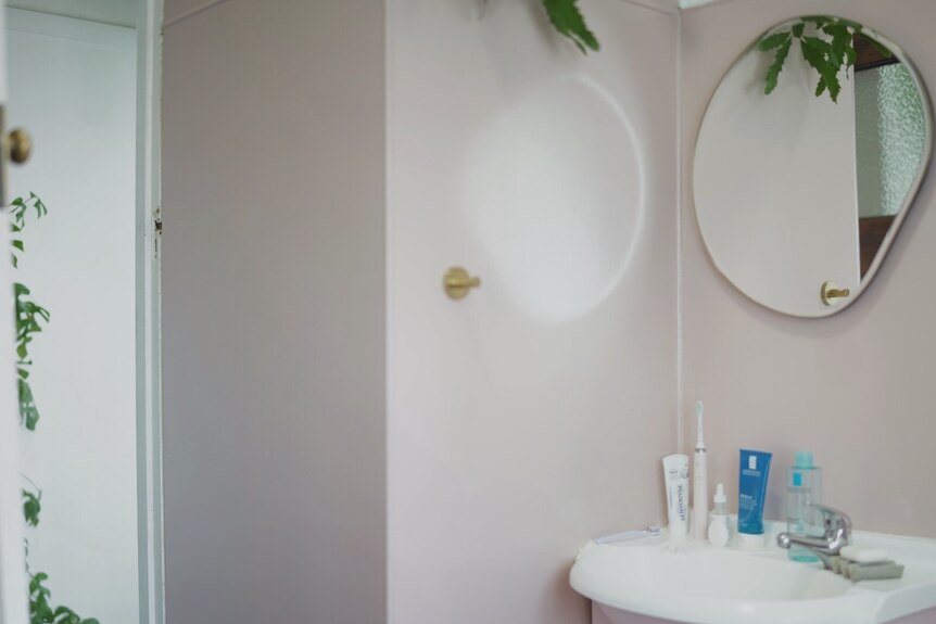 A pink bathroom is seen with a wavy mirror hanging above the wall, a sink with a handful of bathroom products below.