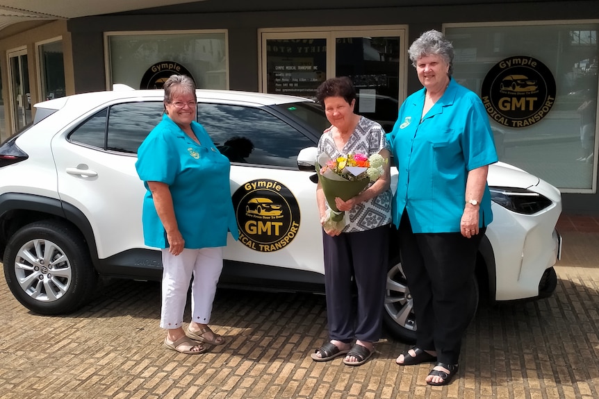 Three women stand in front of white car smiling at camera 