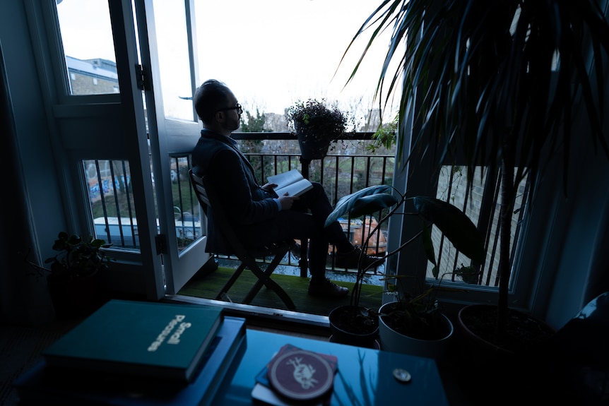A man sits in a chair on a small and dimly-lit balcony, looking out at the world while reading a book