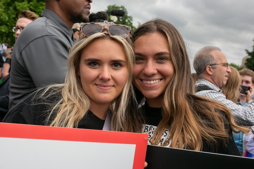 A blonde woman and her brunette friend stand side-by-side at a protest smiling