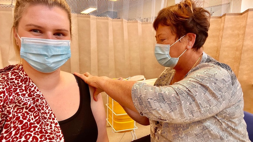 Two women wearing blue face masks, one with her hand on the arm of the other in order to insert a needle
