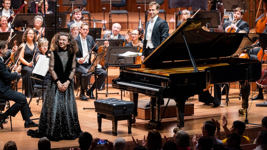 PIanist Marie-Ange Nguci standing in front of the Sydney Symphony Orchestra wearing a resplendent wizard-like dress